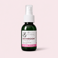 A dark green glass spray bottle of Bojuna Bathroom Spray with a white pump dispenser and a white label featuring pink and black text, set against a solid pale pink background.