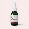 A dark green glass bottle of Botuna Bathroom Spray in Citrus Spice scent, featuring a white spray nozzle and a white label with brown accents, set against a solid pale pink background.