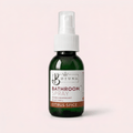 A dark green glass bottle of Botuna Bathroom Spray in Citrus Spice scent, featuring a white spray nozzle and a white label with brown accents, set against a solid pale pink background.