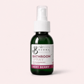 A dark green glass spray bottle of Bojuna Bathroom Spray, Very Berry, with a white pump and a white label, set against a solid pale pink background.