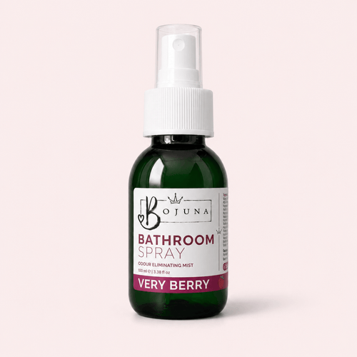 A dark green glass spray bottle of Bojuna Bathroom Spray, Very Berry, with a white pump and a white label, set against a solid pale pink background.
