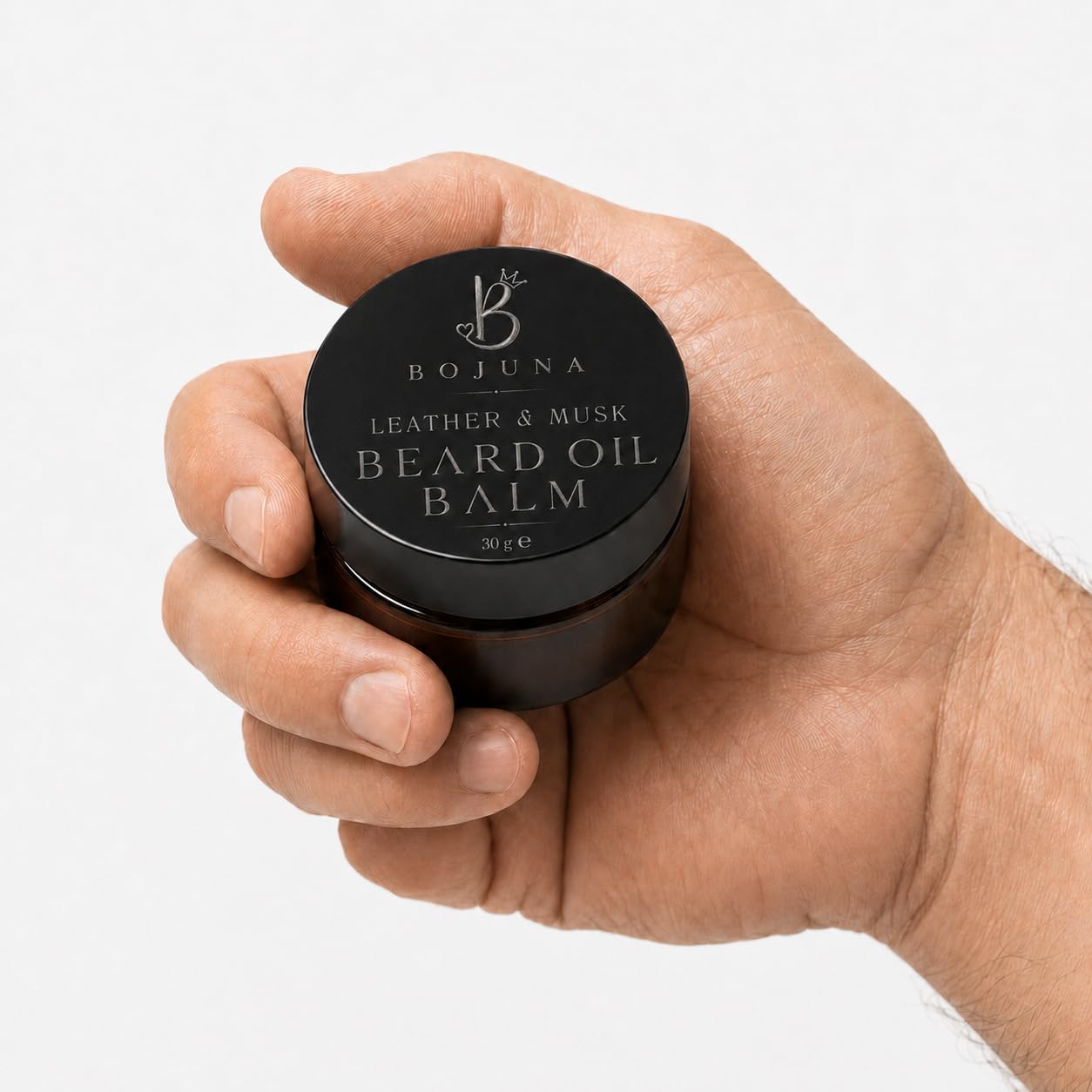 A hand holding a small black round jar of Bojuna Leather & Musk Beard Oil Balm against a clean white background.