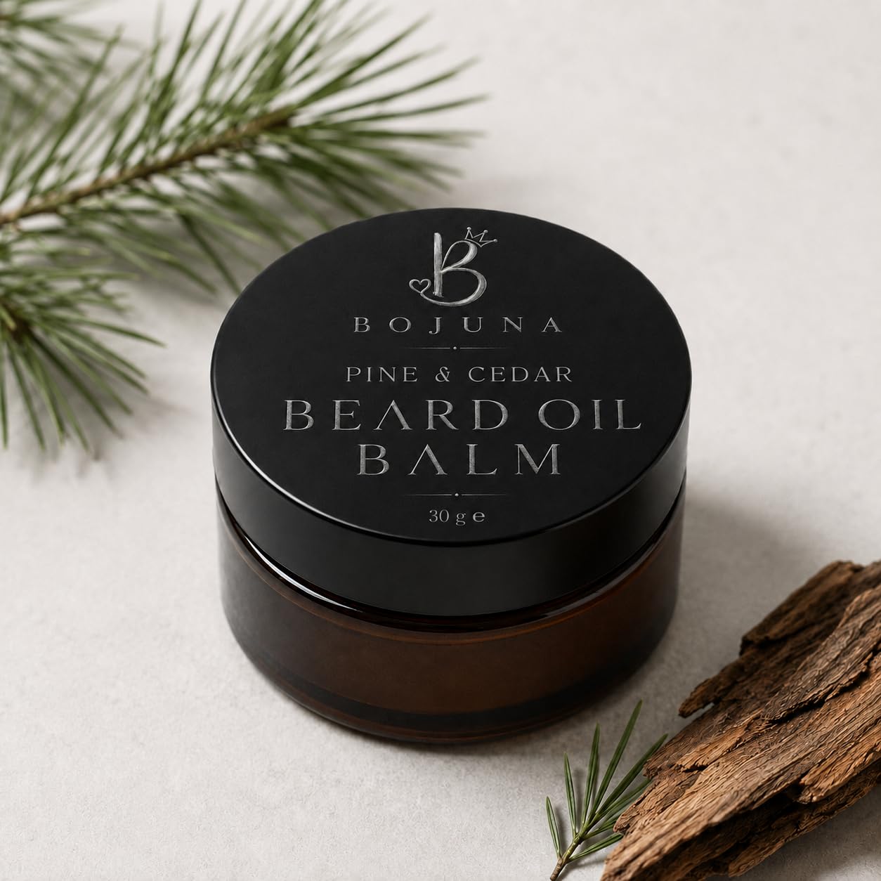 A jar of Bojuna Pine & Cedar Beard Oil Balm, 30g, in a dark amber glass jar with a black lid, styled with pine needles and wood bark on a light gray background.