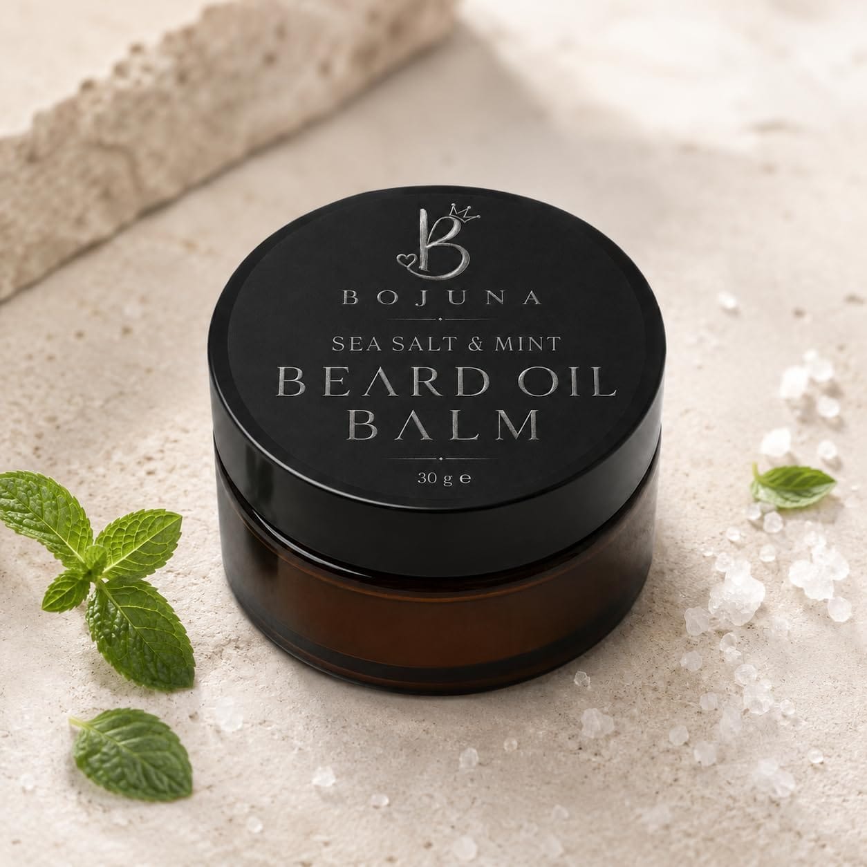 A jar of Bojuna Sea Salt & Mint Beard Oil Balm sits on a light stone surface with mint leaves and sea salt crystals around it. The product has a black lid with silver lettering and a dark amber glass jar.