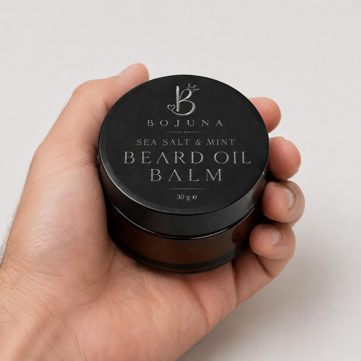 A hand holding a round black jar of Bojuna Sea Salt & Mint Beard Oil Balm against a light gray background.