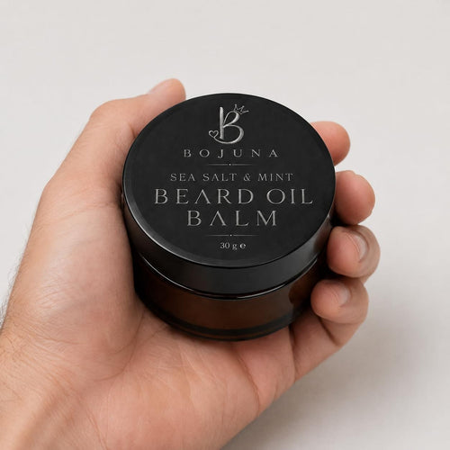 A hand holding a round black jar of Bojuna Sea Salt & Mint Beard Oil Balm against a light gray background.