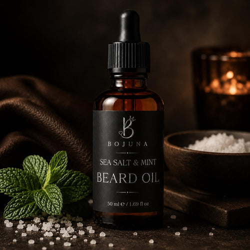 A bottle of Bojuna Sea Salt & Mint Beard Oil in an amber glass dropper bottle with a black label and black cap, styled on a dark rustic surface with mint leaves, coarse salt, and a warm candle in the background.