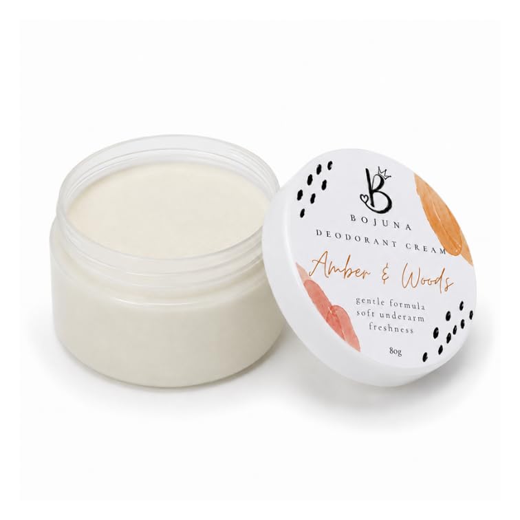 An open clear plastic jar of Bojuna Deodorant Cream next to its white lid, which features a minimalist design with black polka dots and orange and pink botanical illustrations. The cream inside is a warm off-white color, and the lid is labeled 'Amber & Woods'.