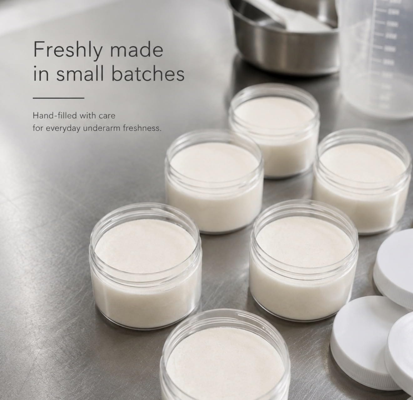 Several small clear jars filled with a pale off-white cream or balm are arranged on a stainless steel surface, with one jar open and its white lid placed beside it. Text on the left reads, 'Freshly made in small batches' and 'Hand-filled with care for everyday underarm freshness.