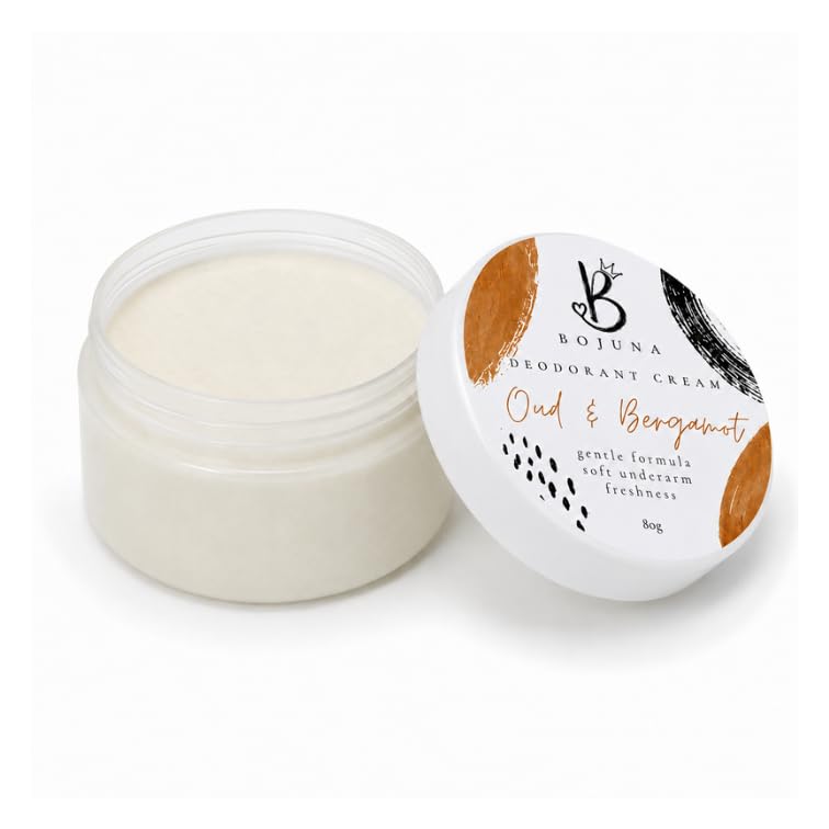 An open clear plastic jar of Bojuna Oud & Bergamot Deodorant Cream next to its white lid, which features gold and black graphic accents, on a clean white background.