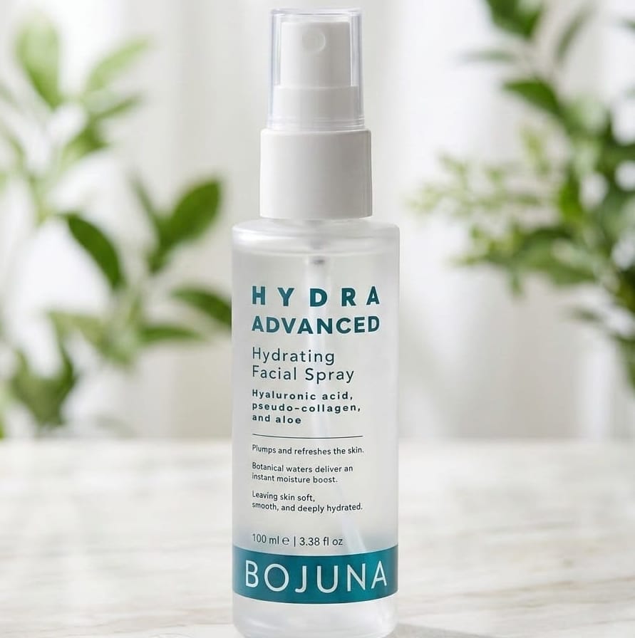 A clear plastic spray bottle of BOJUNA Hydra Advanced Hydrating Facial Spray with a white pump, set on a light marble surface with a soft-focus background of green leaves and white curtains.