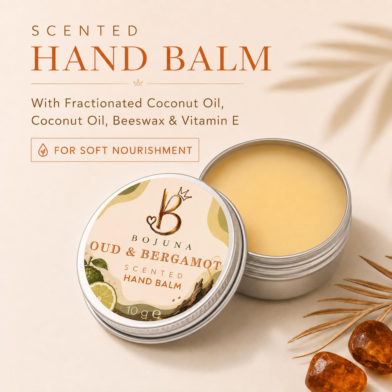 An open round tin of Bojuna Oud & Bergamot scented hand balm sits beside its labeled lid on a warm cream background with decorative dried leaves and amber-like stones. Text on the image lists ingredients including fractionated coconut oil, coconut oil, beeswax, and vitamin E.