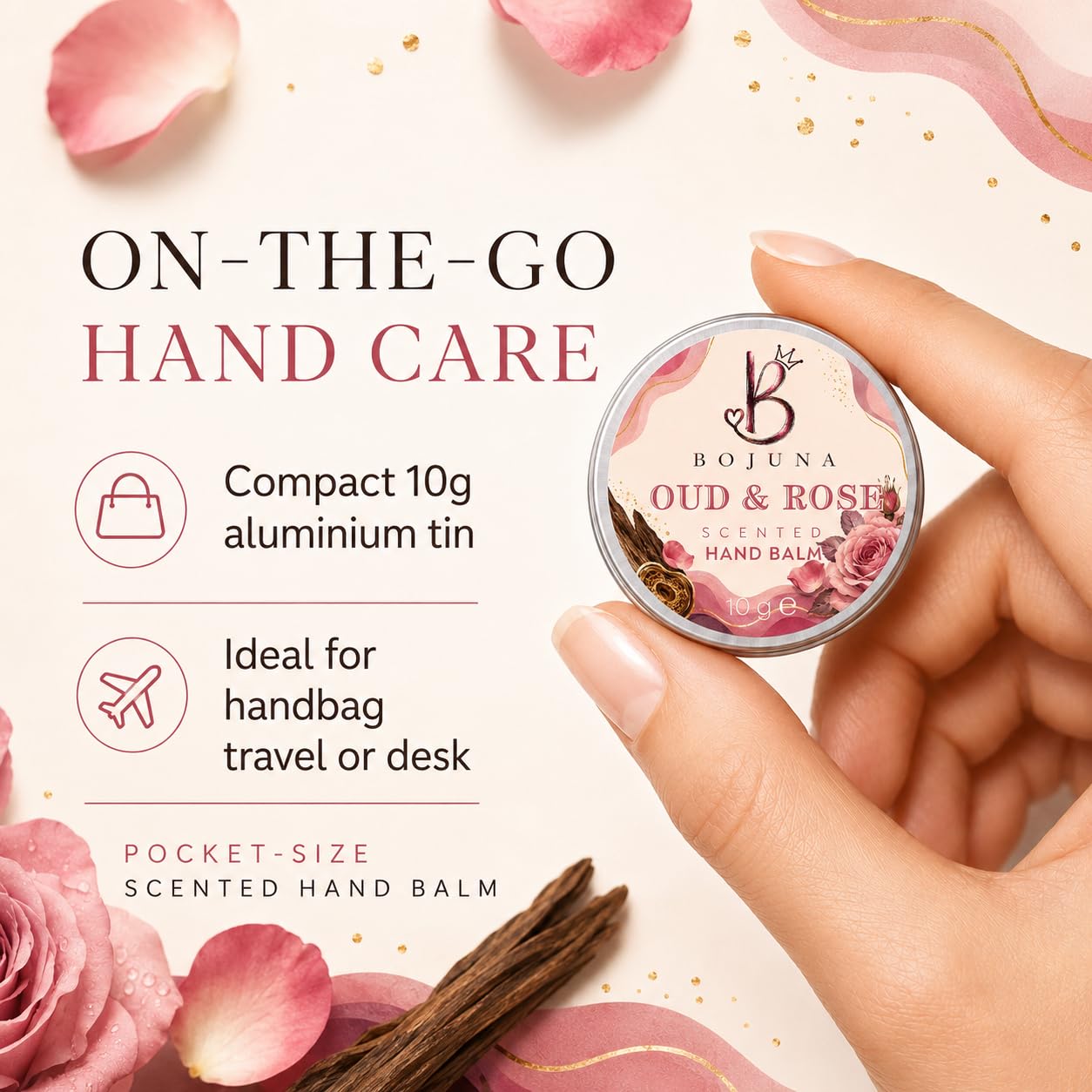 A hand holds a small round tin of Bojuna Oud & Rose Scented Hand Balm against a soft cream background with pink rose petals, gold speckles, and text describing it as on-the-go hand care.