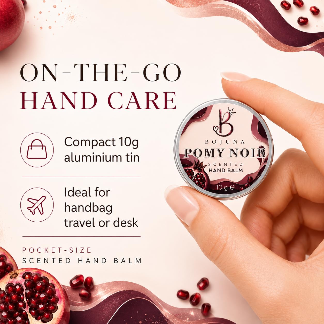 A hand holds a small round tin of Bojuna Pomy Noir Scented Hand Balm against a light pink background with promotional text and pomegranate graphics.