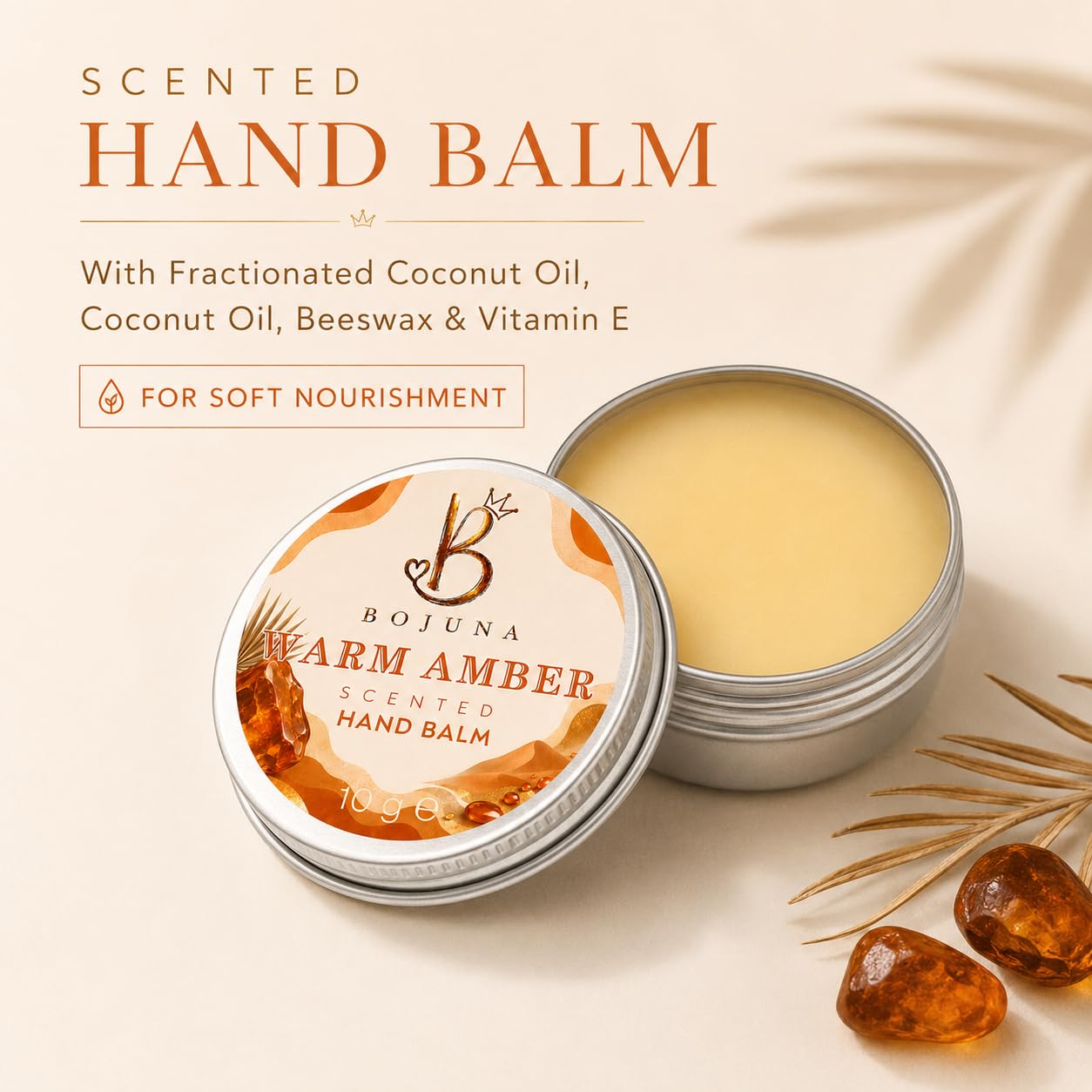 Bojuna Warm Amber Scented Hand Balm shown in two silver round metal tins on a warm beige background with decorative palm leaves and amber stones. Text on the image lists ingredients including coconut oil, beeswax, and vitamin E.