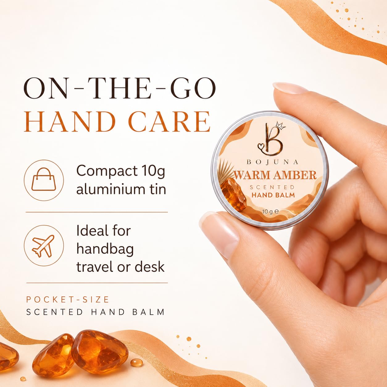 A hand holds a small round tin of Bojuna Warm Amber Scented Hand Balm against a clean cream background with product text and decorative amber droplets.