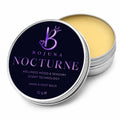 An open round silver tin of Bojuna Nocturne Hand & Foot Balm, showing the pale yellow balm inside next to its dark navy blue lid with purple text and logo.