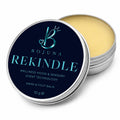 An open round metal tin of Bojuna Rekindle hand and foot balm, showing the pale yellow balm inside, with the lid leaning in front displaying a dark navy label with light blue text.