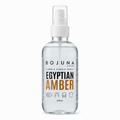 A clear plastic spray bottle of Bojuna Home Egyptian Amber Linen & Fabric Spray, featuring a white spray nozzle and a label with black and orange text, set against a plain white background.